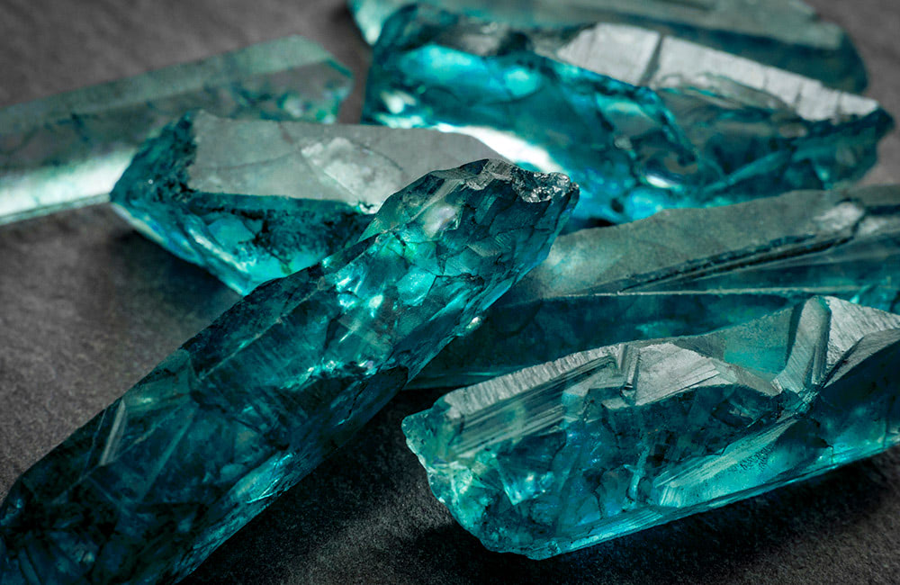 Aquamarine crystals lie scattered on a dark surface, each displaying unique internal fractures and facets, casting a luminous, almost ethereal glow.