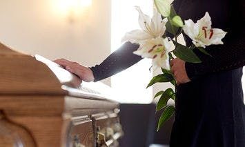 A person holding white lilies stands by a wooden casket with one hand resting on it, in a softly lit room, suggesting a funeral or memorial setting.